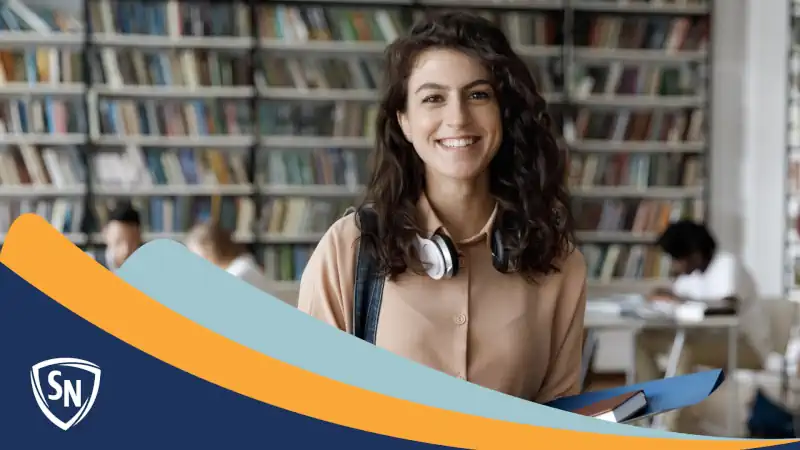 woman in college library holding folder and book, smiling in our direction